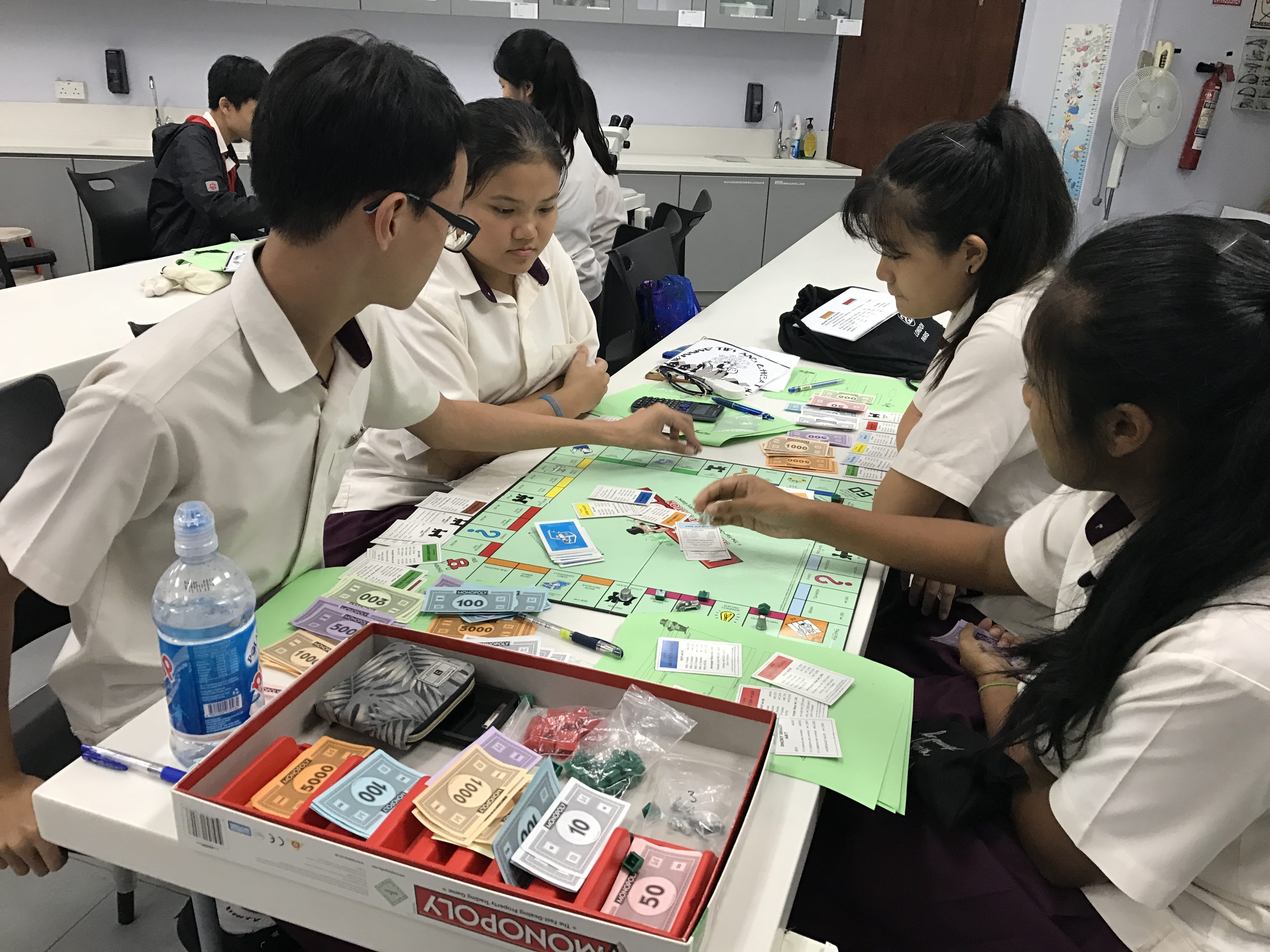 Four students in white uniforms play Monopoly at a classroom table with the game board and money visible.